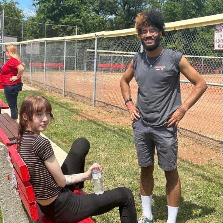 Smiling girl on a bench with a boy standing beside her, both looking at the camera.